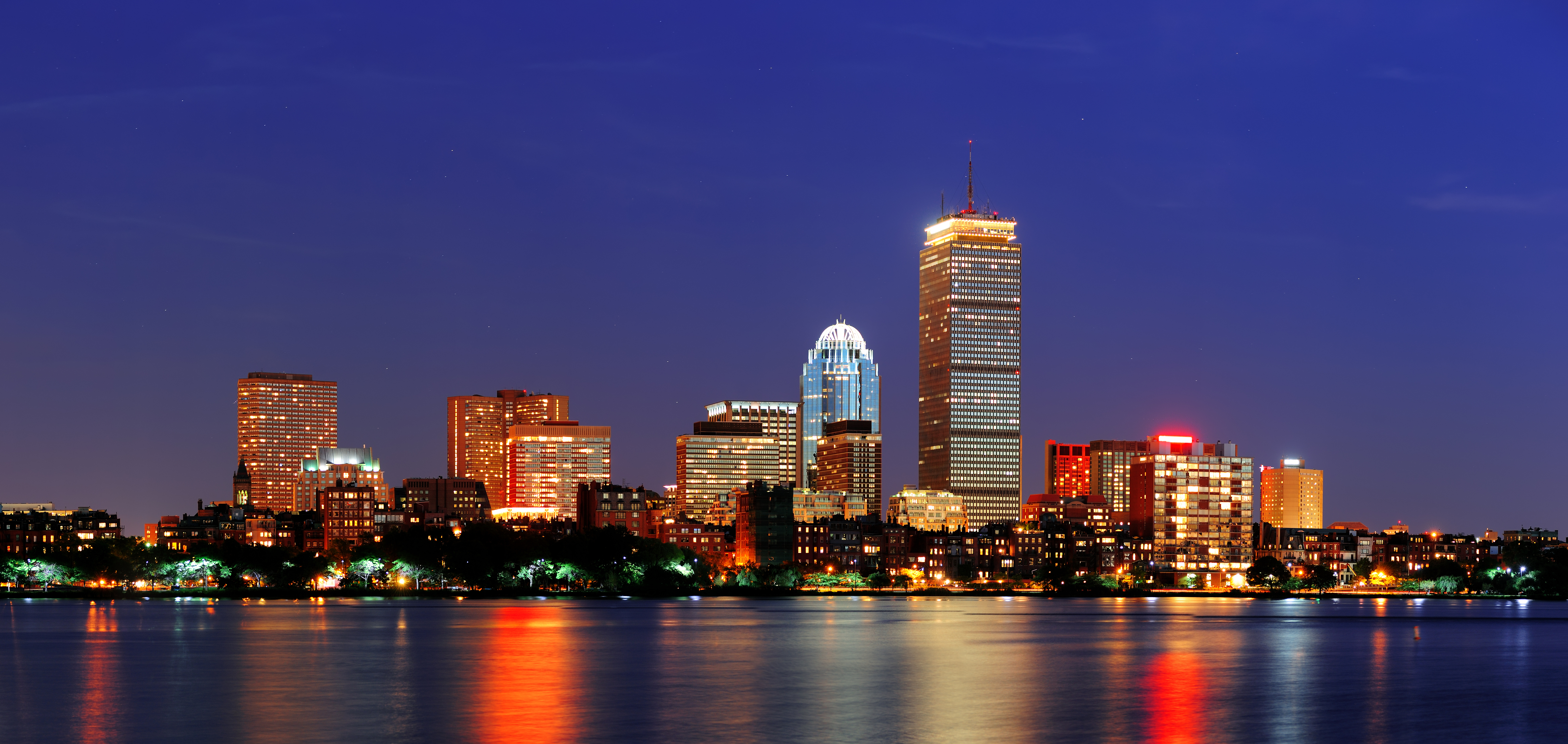City skyline at night with illuminated skyscrapers reflecting on water.