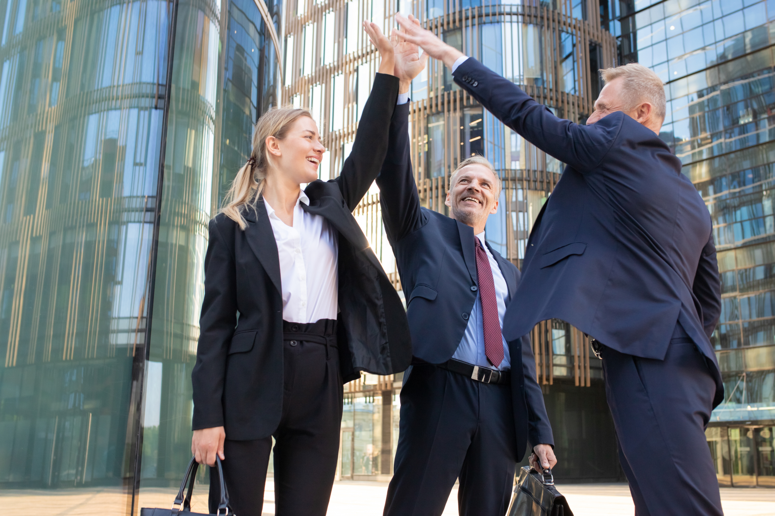 Three cheerful business professionals giving a high five outdoors in front of a modern office building.